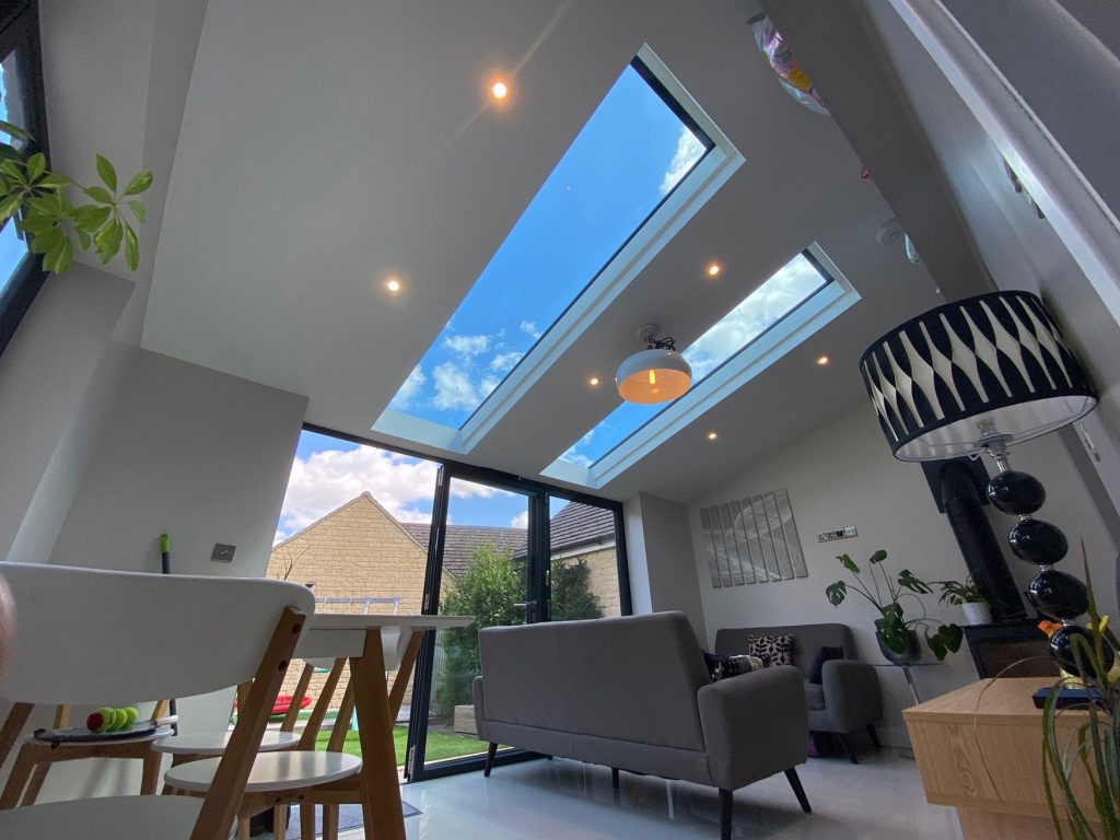 A modern living room with skylights showing a blue sky and clouds. Grey sofas, a black and white lamp, indoor plants, and Cairnview Windows and Doors frame the garden and houses outside through large glass doors.