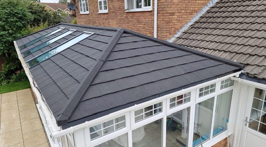 A conservatory with a dark grey, tiled pitched roof featuring skylights by Cairnview Windows and Doors, attached to a brick house with white-framed windows and uPVC doors Perth, with a garden area beside a paved patio.