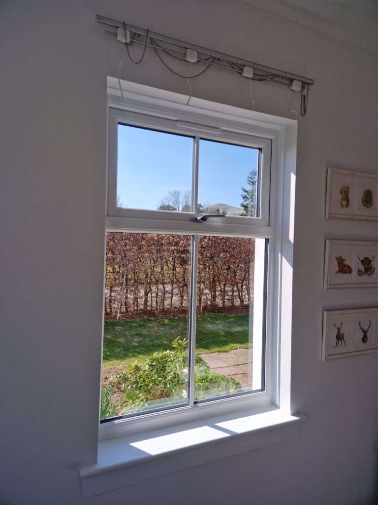 A sunny garden with green grass and a hedge is visible through a white-framed uPVC window perth. The window is partly open and sunlight casts shadows on the interior white wall, which displays framed artwork.