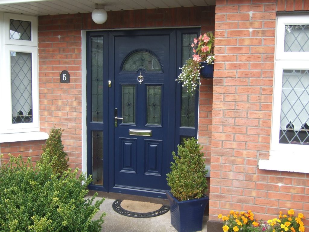 A dark blue uPVC door with decorative glass panels, flanked by tall potted evergreens and hanging flowers, set in a red brick house with white-trimmed windows from Cairnview Windows and Doors and a black house number 5 plaque.