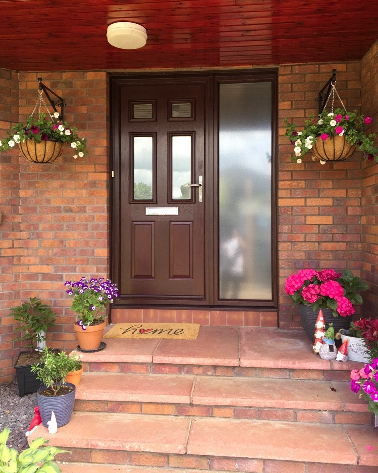 A brown uPVC door with frosted glass panel is framed by hanging flower baskets and potted plants on brick steps. A doormat reading Home lies at the entrance, next to a small garden gnome and beneath charming windows.