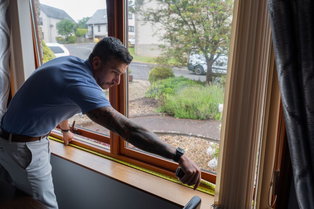 A man with tattoos, wearing a blue shirt and white pants, measures a large wooden window frame using a yellow tape measure in a bright room that overlooks a garden and street—perfect for installing new uPVC windows Perth.
