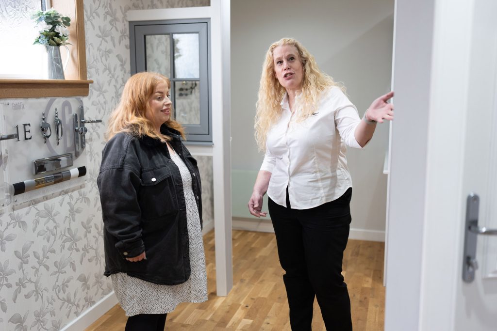 Two women stand indoors, chatting in a hallway near Cairnview Windows and Doors. One, with curly blonde hair and a white shirt, gestures as she speaks to the red-haired woman in a black jacket. The stylish doors hint at their Perthshire location.