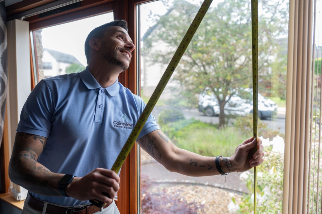 A man in a blue polo shirt measures a window with a tape measure, smiling as he works inside a house. Trees and parked cars are visible outside through the window, showcasing quality windows from Cairnview Windows and Doors.