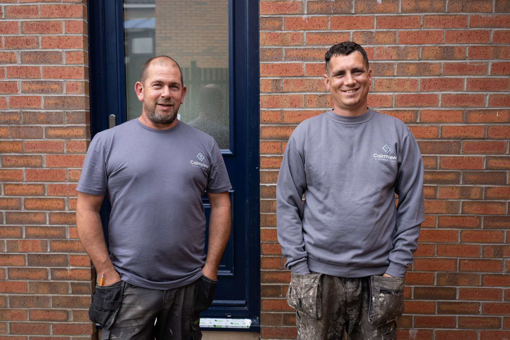 Two men wearing grey work shirts and construction pants stand smiling in front of a brick wall and a blue door, representing Cairnview Windows and Doors—specialists in uPVC doors Perth with tools and building materials visible on their pants.