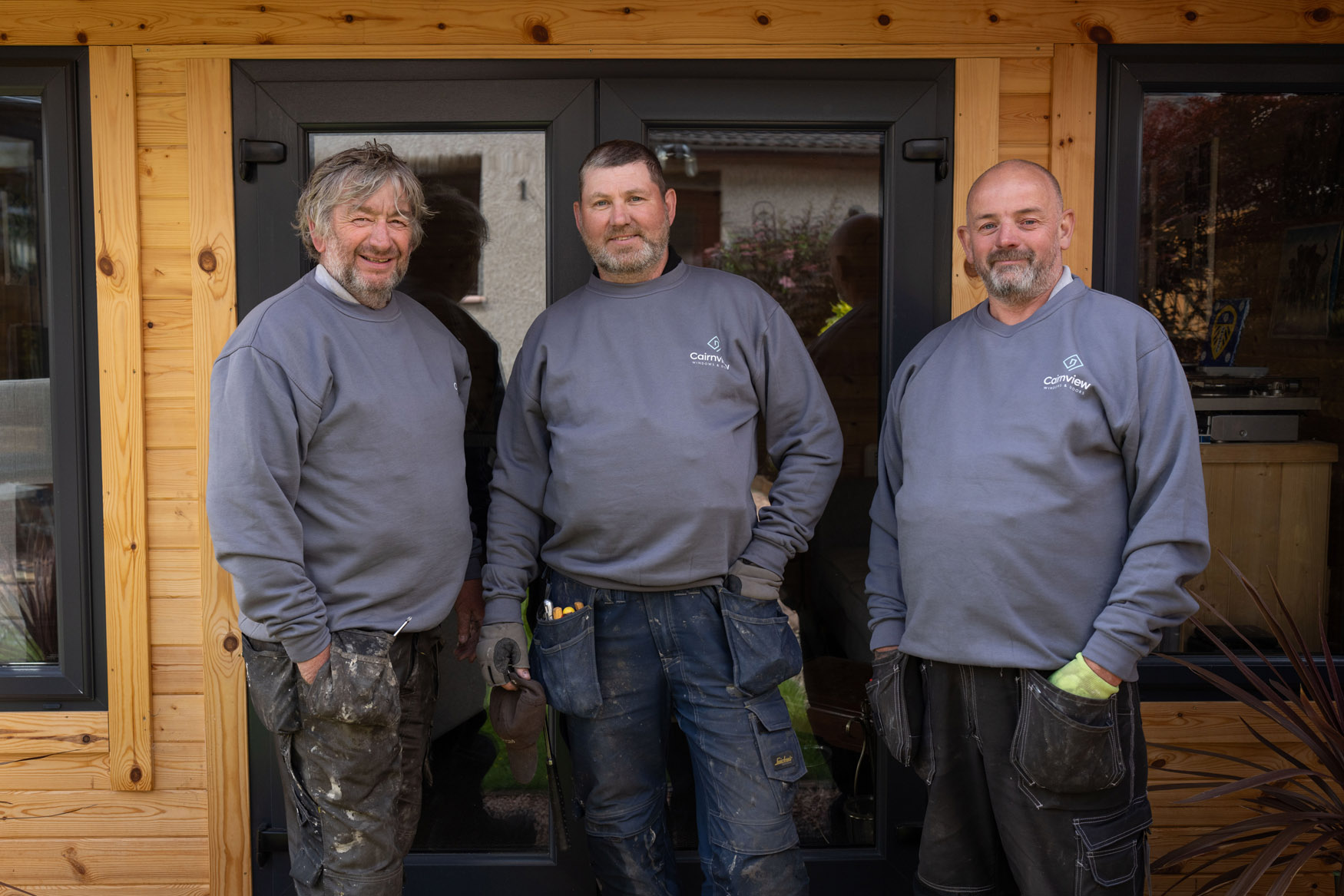 Three men wearing matching gray sweatshirts and work pants stand in front of a wooden building with glass doors, smiling and posing for the camera—showcasing quality doors perfect for any Perthshire home.