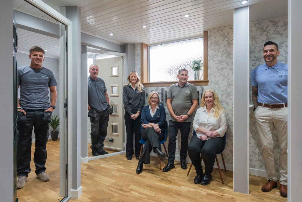 Seven people, four men and three women, are posing and smiling in a modern office space with wood floors, white walls, uPVC windows, and a floral accent wall. Some are seated while others stand, creating a welcoming atmosphere.