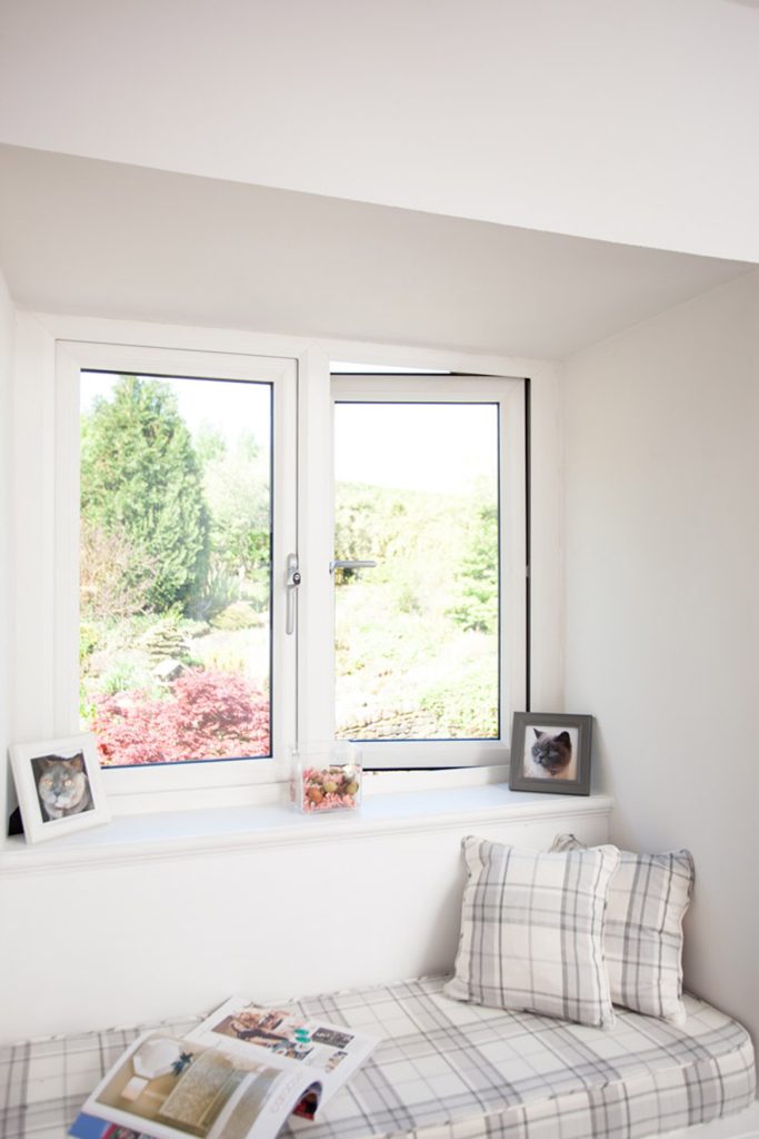 A sunny window nook with two framed cat photos on the sill, plaid cushions on a bench, a magazine, and a jar, overlooking a lush garden through bright uPVC windows Perth residents love.