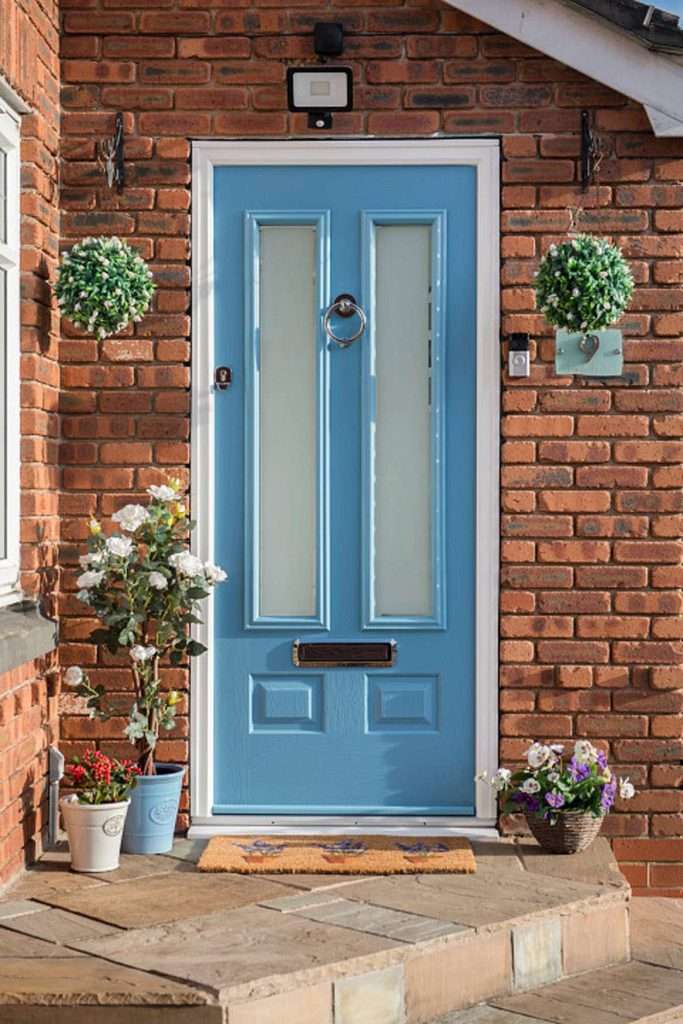 A light blue front door with frosted glass panels set in a red brick wall, surrounded by potted flowers and hanging plants, with a welcome mat on the stone doorstep.