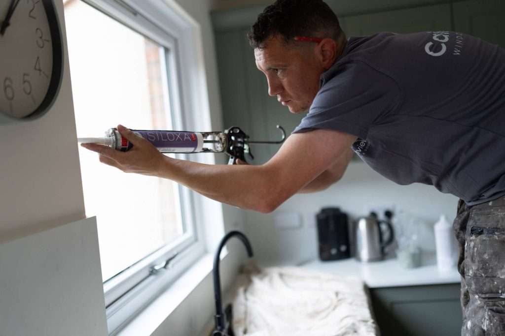 A man in work clothes applies sealant around a kitchen window using a caulking gun. The kitchen counter below is covered with a sheet, and various appliances are visible in the background.