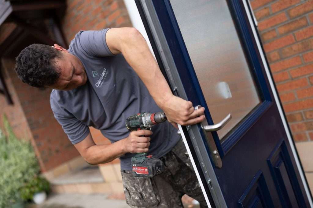 A man wearing a gray t-shirt uses a power drill to install or repair a blue door on a brick house.