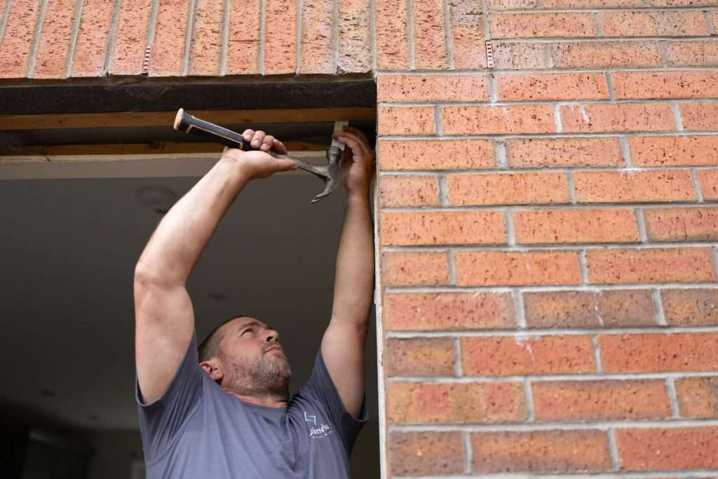 A man in a gray shirt uses a hammer to work on the wooden frame of a doorway in a brick building, reaching up with both hands.