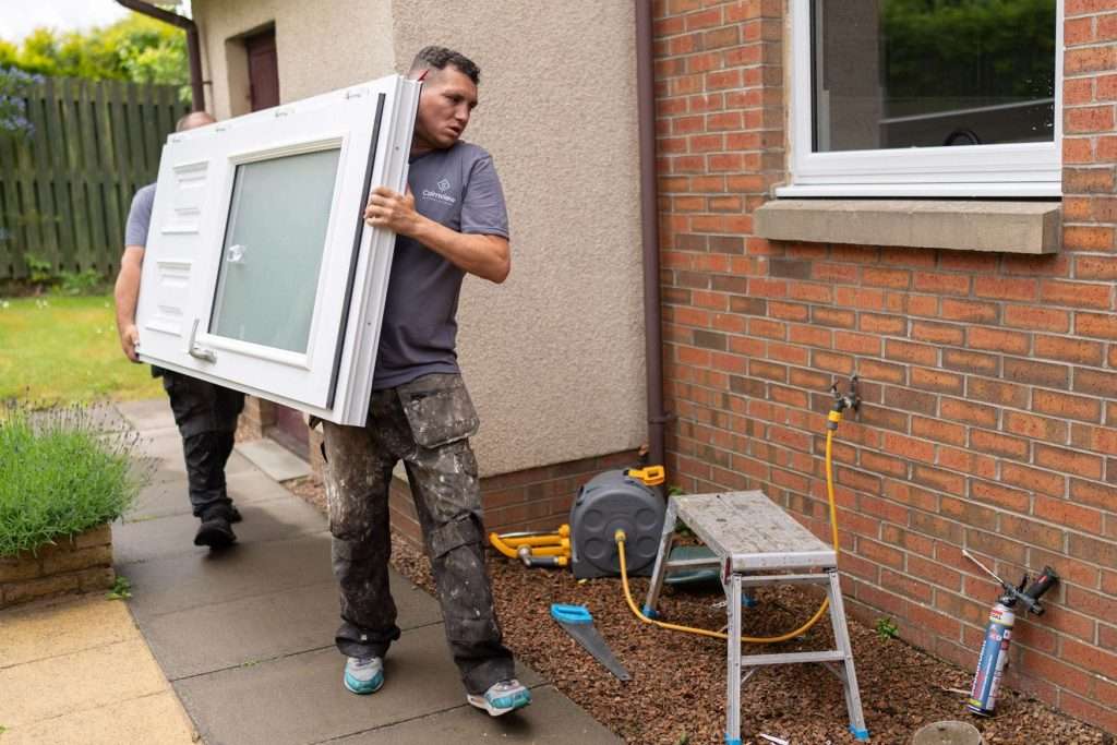 A man in work clothes carries a window frame outside a brick house. Tools, a hose reel, and a small step ladder are on the ground nearby. Another person is partially visible behind him.