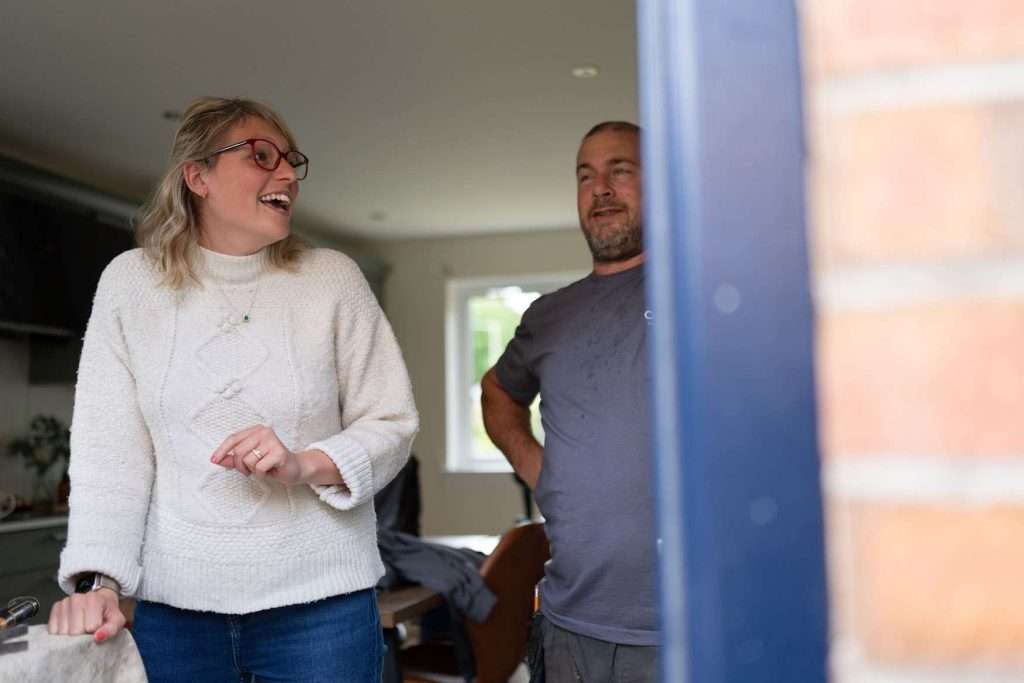 A woman in a white sweater and glasses laughs while standing next to a man in a gray shirt. They are indoors, near a window, with natural light coming in. The scene is casual and cheerful.