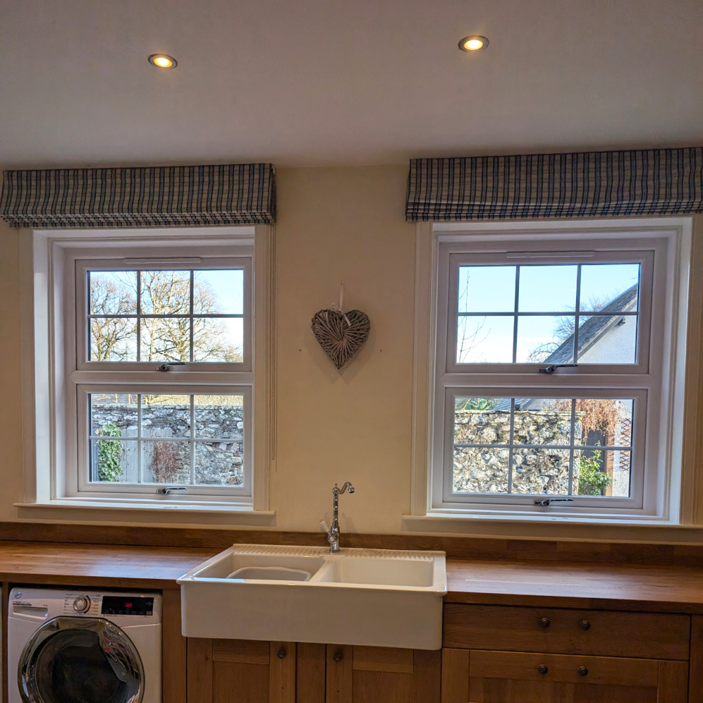 A kitchen with two windows above a white farmhouse sink, wooden countertops, and a washing machine on the left. A heart-shaped decoration hangs between the windows, with a stone wall and trees visible outside.