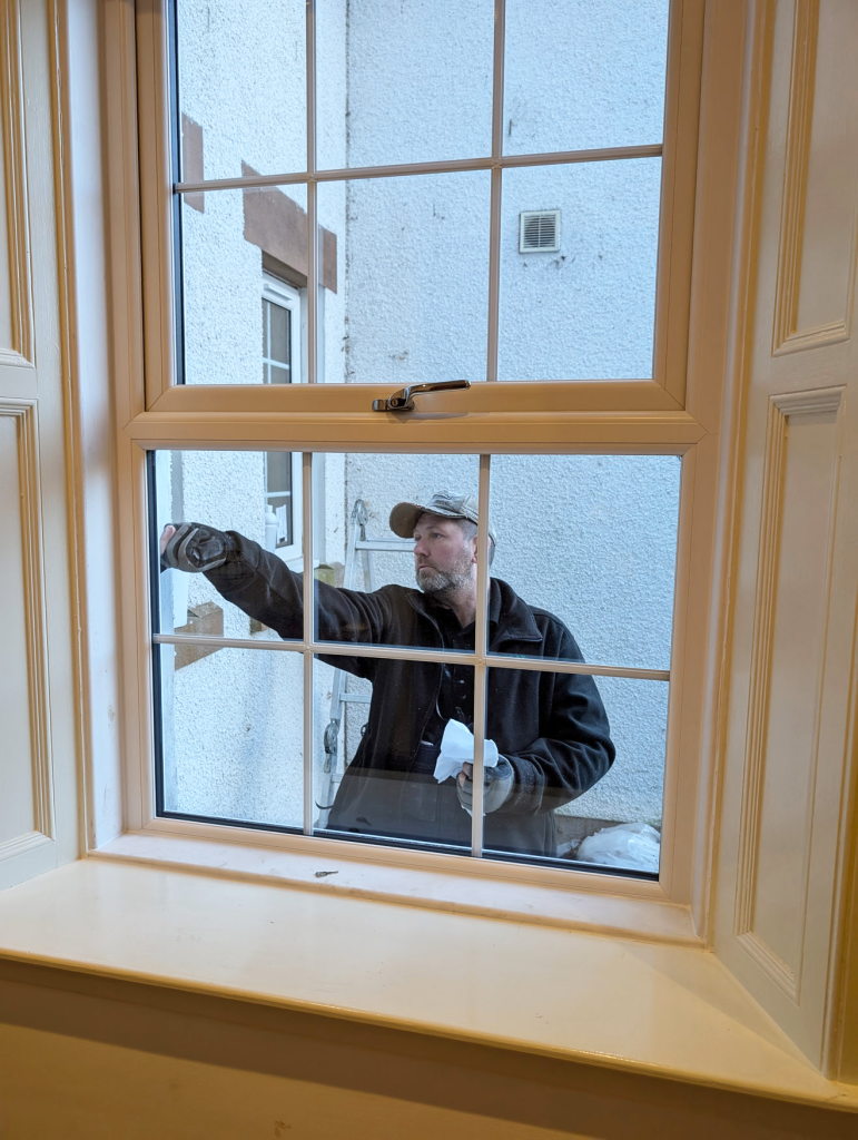 A man wearing a cap and dark jacket is painting or repairing the exterior window frame of a building, as seen through a window from inside the room.