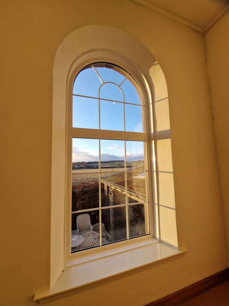 A sunlit arched window with white trim overlooks a patio with white chairs and a table, and a scenic landscape of fields and distant hills under a clear blue sky.