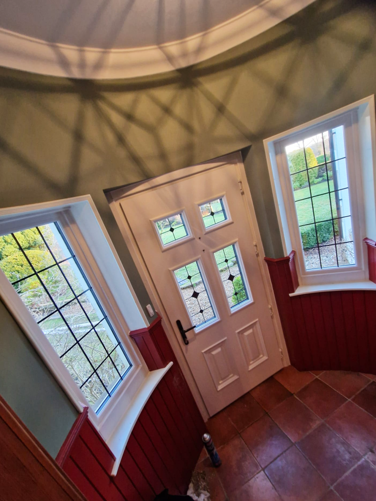 A white front door with four glass panels is flanked by two large windows with a similar grid design. The entryway walls are green and red, with a tiled floor and a garden visible outside. Shadows from a light fixture appear on the ceiling.