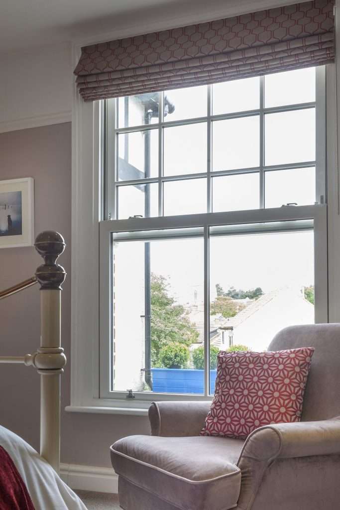 A cozy bedroom corner with a light purple armchair and a red patterned cushion by a large window, offering a view of trees and houses outside. A bed with a red throw is partially visible in the foreground.