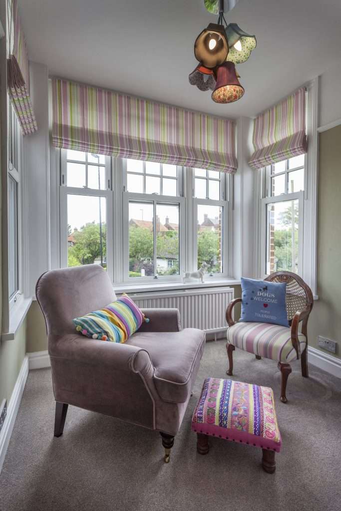 A cozy reading nook with large windows, a plush armchair, a colorful striped pillow, a matching footstool, a vintage chair, and vibrant striped Roman blinds overlooking a green, leafy neighborhood.