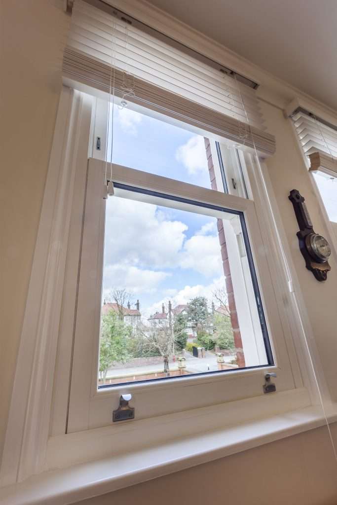 A white-framed window partially open, with blinds above and a view of trees, houses, and a cloudy blue sky outside. A vintage wall clock hangs to the right of the window.