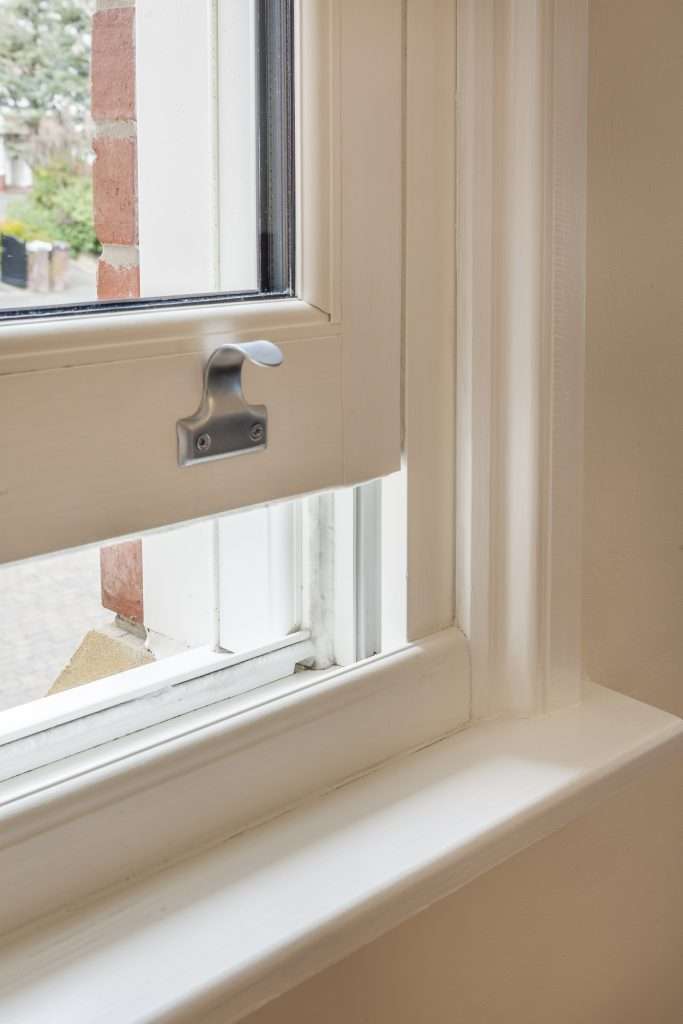A close-up view of a white, open sash window with a metal latch in a house, showing part of the exterior brick wall and blurred greenery outside.