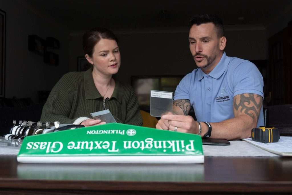 A man and woman sit at a table, reviewing glass samples. The man wears a blue polo and has tattoos; the woman wears a green cardigan. A green Pilkington Textured Glass sample book is open in front of them.