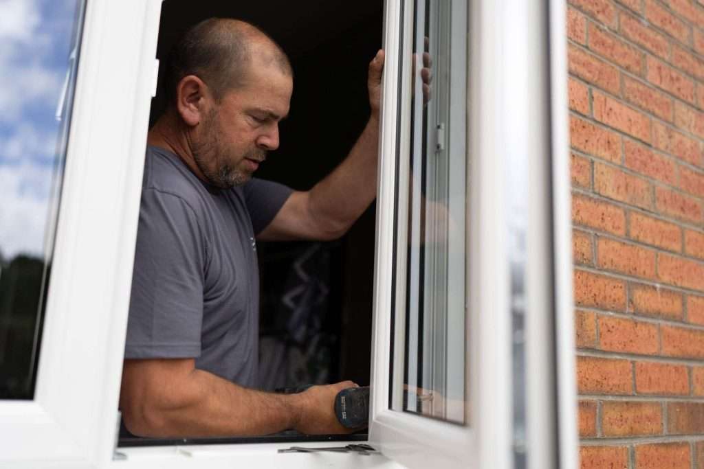 A man in a gray shirt installs or repairs a white-framed window, holding a power drill, with a red brick wall visible beside him.