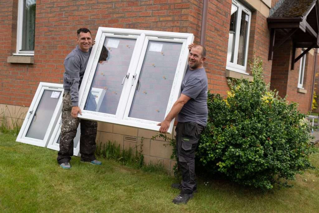 Two men are smiling while carrying a large window frame in front of a brick house with green grass and a bush nearby. Another window frame is propped against the house behind them.