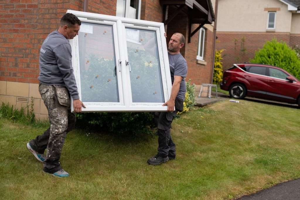 Two men carry a large white-framed window outside a brick house with a garden. Both wear gray shirts and work pants. A red car is parked on the driveway in the background.