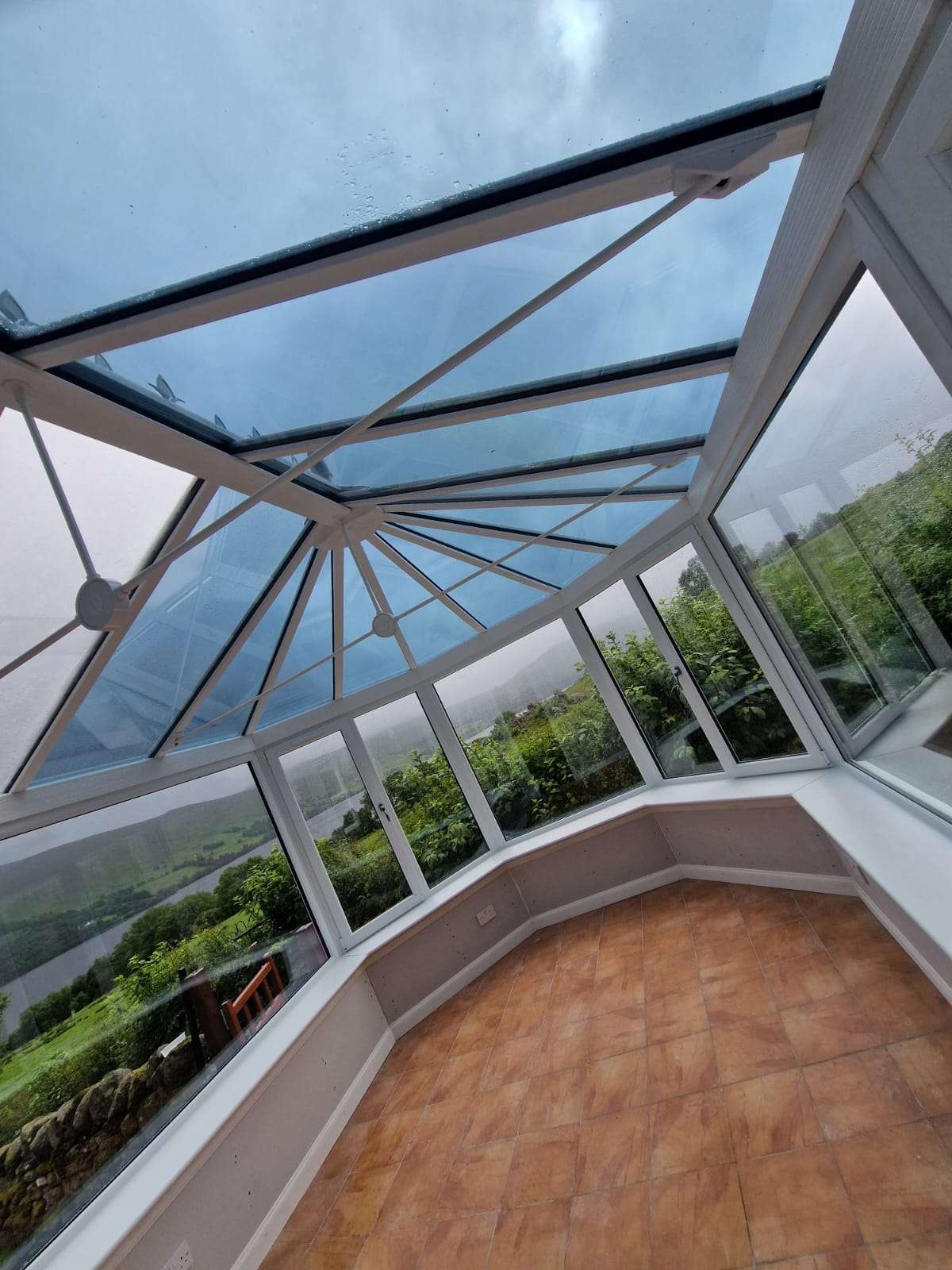 A bright conservatory replacement in Aberfeldy featuring a glass roof, large windows, tiled floor, and views of green landscape and a river outside under a cloudy sky.