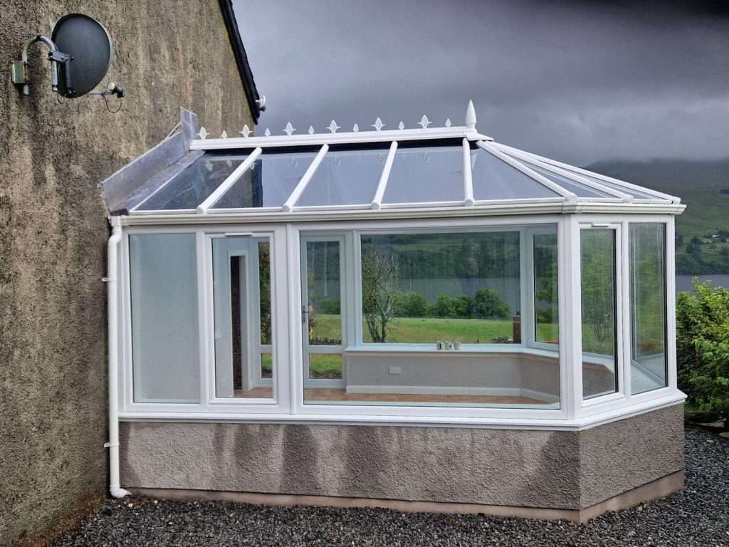 A white-framed glass conservatory replacement in Aberfeldy is attached to a stone house, featuring large windows and a slanted glass roof. The background reveals a cloudy sky and green hills with a shimmering body of water.