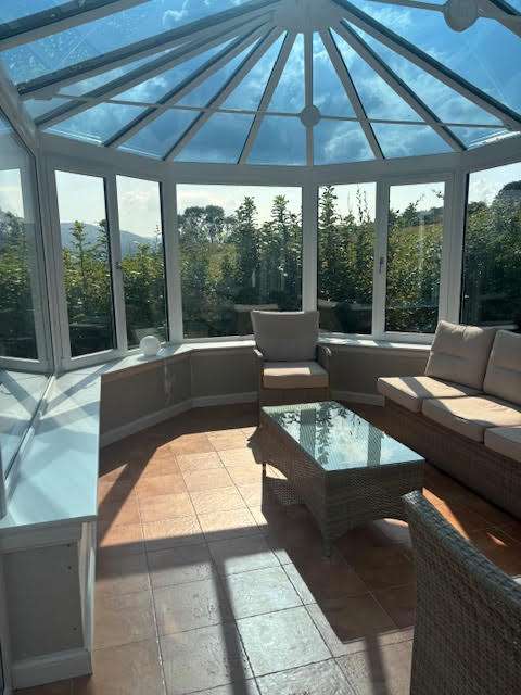 A bright conservatory with glass walls and ceiling, furnished with a beige cushioned sofa, an armchair, and a glass-top wicker table. Sunlight streams in, casting shadows on the tiled floor. Shrubs and trees are visible outside.