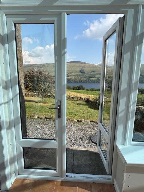 A glass door is open, revealing a garden with a gravel path, green grass, and rolling hills in the distance under a blue sky with scattered clouds.
