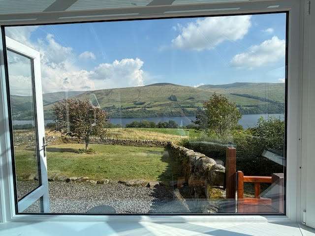 View through a large window showing a grassy yard, stone wall, trees, and a serene lake with green hills and blue sky with clouds in the background. Part of an open glass door and porch railing are visible.