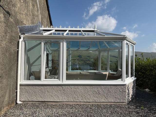 A white-framed glass conservatory is attached to a stone building, with clear blue skies and distant hills visible through the windows. The conservatory sits on a gravel area and contains light-colored furniture.