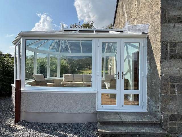 A modern glass conservatory attached to a stone house, featuring large windows, a white frame, and several beige cushioned chairs inside. Sunlight streams in, and greenery is visible through the windows.