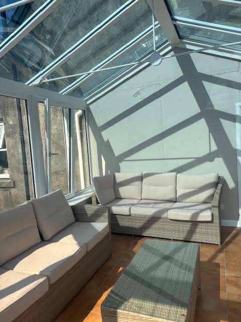 A sunlit conservatory with glass walls and ceiling, featuring two beige cushioned sofas and a woven coffee table on a wooden floor. Sunlight casts geometric shadows across the space.