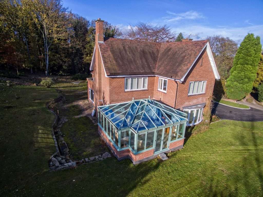 Aerial view of a two-story red-brick house with a glass conservatory attached, surrounded by a grassy yard, trees, and a curved driveway under a blue sky.