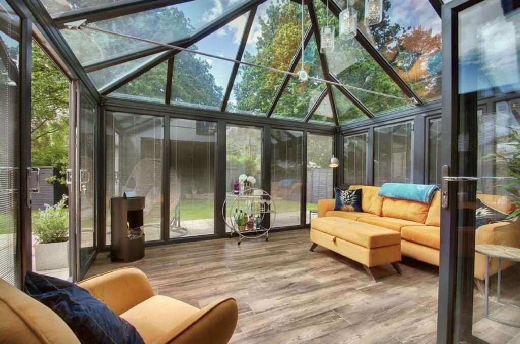 A bright sunroom with glass walls and ceiling, furnished with a yellow sofa, matching armchair, small black stove, and a bar cart. The room overlooks a green garden with trees outside.