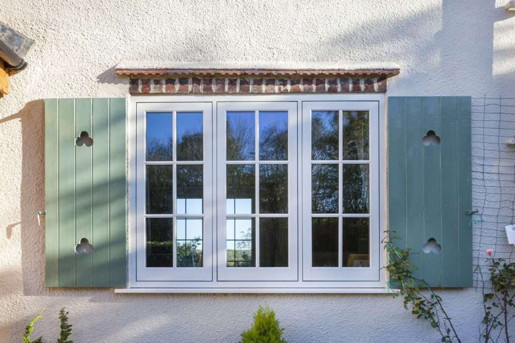 A white-framed window with green wooden shutters on both sides, set in a light-colored stucco wall, reflects trees and sky in its glass panes. Some plants grow below the window.