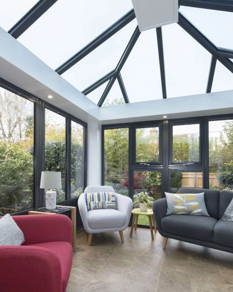 A bright conservatory with a glass roof by Conservatory Roofs Dunkeld, featuring black-framed windows, a red armchair, a gray sofa, a white armchair, a small lamp, and decorative cushions. Lush greenery is visible outside.