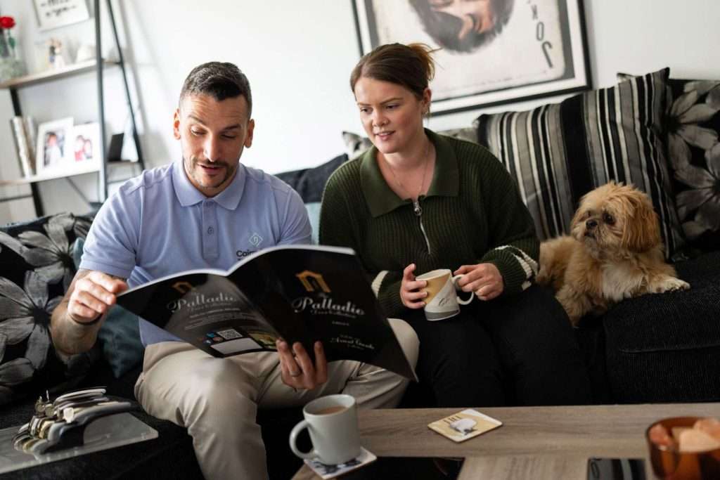A man and woman sit on a couch together, looking at a magazine about visiting windows doors showroom while holding mugs. A small dog sits beside them on the couch. The scene appears relaxed and cosy in a home living room.