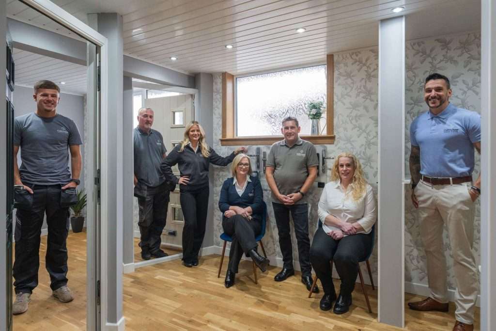 Seven people pose together in a modern, well-lit office space with wooden floors and patterned wallpaper, as if visiting a windows and doors showroom. Four stand while three are seated, all smiling and dressed in business or casual attire.