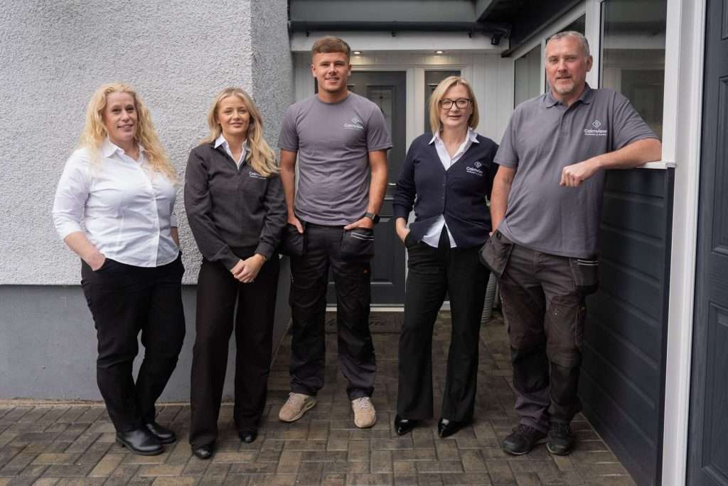 Five people stand outside a building, smiling for a group photo. Visiting a windows and doors showroom, two men in grey work shirts and three women in business attire pose on a paved area beside a house with grey and white walls.