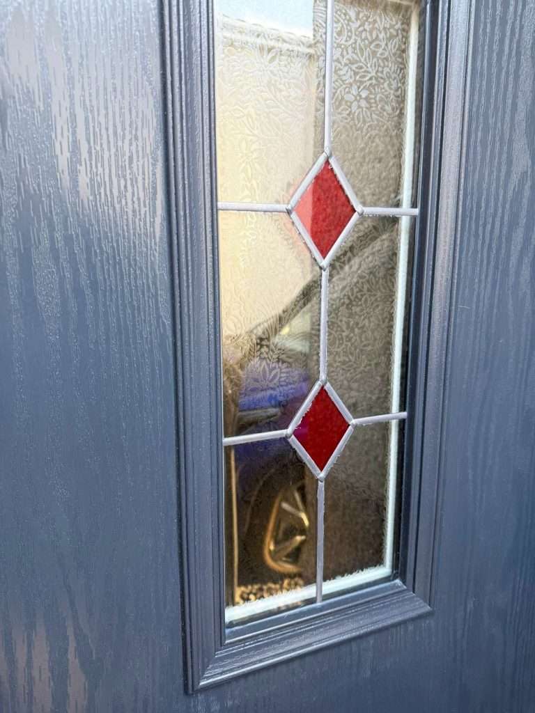 Close-up of a grey textured door with a frosted glass window panel featuring a decorative pattern and two red diamond shapes—perfect inspiration for house renovations Perth homeowners will love. Through the glass, part of a car wheel is visible outside.