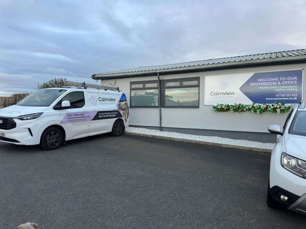 A white van and car are parked outside a gray building with signs for Cairnview. A flower box sits under the window, and a large sign welcomes visitors to the windows and doors showroom and office.