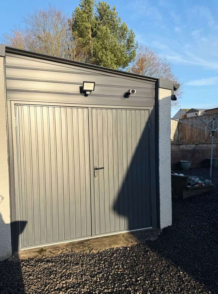 A modern gray metal garage with double doors, security camera, and outdoor light fixture is perfect for house renovations Perth; gravel ground and garden area sit beside the building, framed by trees and blue sky in the background.