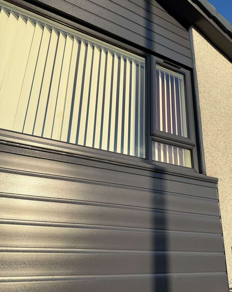 Sunlight casts a shadow on a modern building exterior with gray metal siding and a window covered by vertical white blinds. The blinds are partially open, letting in some light.